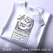 Футболка золоті руки, біла, складена на поверхні, фон світло-сірий, принт чорного кольору з написами та малюнками, включаючи фрази 'Чиста душа' та 'Золоті руки', з зеленим ярликом.