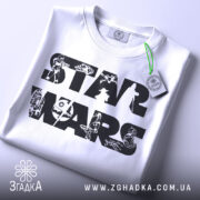 Футболка зоряні війни білого кольору, складена на столі, видно етикетку з зеленим шнурком, фон світло-сірий, принт з написом 'STAR WARS' та малюнками персонажів і техніки з всесвіту Зоряних війн.