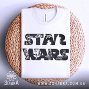 Футболка зоряні війни білого кольору, складена на плетеній кошику, фон світло-сірий, принт з написом 'STAR WARS' та малюнками персонажів і техніки з всесвіту Зоряних війн.