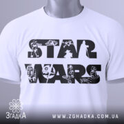 Футболка зоряні війни білого кольору, складена на столі, видно етикетку з зеленим шнурком, фон світло-сірий, принт з написом 'STAR WARS' та малюнками персонажів і техніки з всесвіту Зоряних війн.