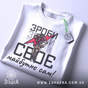 Футболка зроби сам, білого кольору, зображена зверху, на ній видно етикетку з зеленим шнурком, фон світло-сірий, принт аналогічний попередньому, текст чорного кольору.