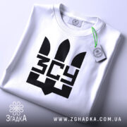 Футболка ЗСУ білого кольору, зображена зверху, складена на столі, фон світло-сірий, принт чорного кольору з тризубом та написом 'ЗСУ', на комірці є етикетка з зеленим шнурком.