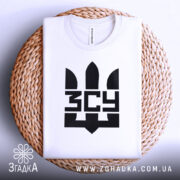 Футболка ЗСУ білого кольору, зображена зверху, складена на плетеному кошику, фон світло-сірий, принт чорного кольору з тризубом та написом 'ЗСУ'.