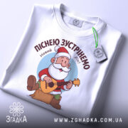 Футболка зустрінемо Новий Рік, біла, складена на столі, фон світло-сірий, принт з дідом Морозом, який грає на гітарі, текст зверху: 'Піснею зустрінемо Новий Рік!', на етикетці зелена стрічка.