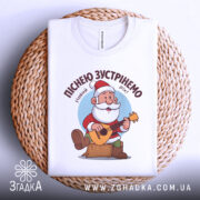 Футболка зустрінемо Новий Рік, біла, складена на плетеній кошику, фон світло-сірий, принт з дідом Морозом, який грає на гітарі, текст зверху: 'Піснею зустрінемо Новий Рік!'