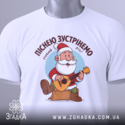 Футболка зустрінемо Новий Рік, біла, складена на столі, фон світло-сірий, принт з дідом Морозом, який грає на гітарі, текст зверху: 'Піснею зустрінемо Новий Рік!', на етикетці зелена стрічка.