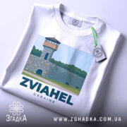 Футболка Звягель місто, біла, складена на столі, фон світлий, принт з малюнком вежі на кам'яній стіні з написом ZVIAHEL UKRAINE, на футболці є етикетка з зеленим шнурком.