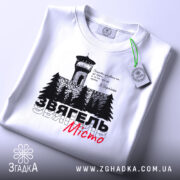 Футболка Звягель білого кольору, зображена на столі, з принтом, що містить вежу та дерева, текст виконаний чорним і червоним кольорами, на футболці є етикетка зеленого кольору.