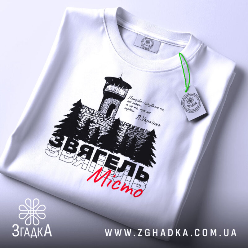 Футболка Звягель білого кольору, зображена на столі, з принтом, що містить вежу та дерева, текст виконаний чорним і червоним кольорами, на футболці є етикетка зеленого кольору.