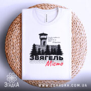 Футболка Звягель білого кольору, зображена на плетеній основі, з принтом, що містить вежу та дерева, текст виконаний чорним і червоним кольорами, фон світло-сірий.