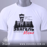 Футболка Звягель білого кольору, зображена на столі, з принтом, що містить вежу та дерева, текст виконаний чорним і червоним кольорами, на футболці є етикетка зеленого кольору.