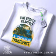 Футболка звичайний тракторист. Біла футболка, складена на плоскій поверхні, з видимим ярликом. Зображення з верхнього ракурсу. Фон світло-сірий. Принт з трактором та текстом: 'Я не блогер, не артист, я - звичайний тракторист' у зеленому та жовтому кольорах.