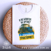 Футболка звичайний тракторист. Біла футболка, складена на плоскій поверхні з плетеного матеріалу. Зображення з верхнього ракурсу. Фон світло-сірий. Принт містить зображення трактора та текст: 'Я не блогер, не артист, я - звичайний тракторист' у зеленому та жовтому кольорах.