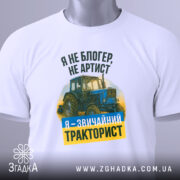 Футболка звичайний тракторист. Біла футболка, складена на плоскій поверхні, з видимим ярликом. Зображення з верхнього ракурсу. Фон світло-сірий. Принт з трактором та текстом: 'Я не блогер, не артист, я - звичайний тракторист' у зеленому та жовтому кольорах.