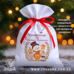 Іменна Торбинка до Миколая на фоні ялинки й вогників, білий габардин 20×30 см з червоною стрічкою та принтом зі сніговиком і звірятками.