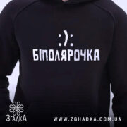 Худі біполярочка чорного кольору, зображене з близької відстані. На передній частині видно принт з написом 'БІПОЛЯРОЧКА' у білому кольорі. Капюшон піднятий, манжети та пояс виглядають зручно.