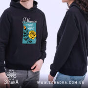 Худі дівчина твоєї мрії, темно-синього кольору, з капюшоном, ракурс з боку, на фоні видно дві людини в худі. Принт з жовтою трояндою та написом на блакитному фоні, є кишеня-«кенгуру», манжети та пояс класичного стилю.