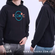 Худі для фанатів космосу. Темно-синій колір, зображення з бокового ракурсу, фон світло-сірий. Два моделі в худі з принтом 'КСМЦ' у кольорах рожевий та блакитний. Видима кишеня-«кенгуру», капюшони на обох моделях, манжети з еластичної тканини.