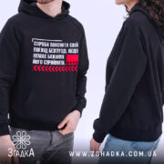 Худі пояснити погляд. Чорний колір, ракурс зліва, фон світлий. Принт на грудях з текстом та червоним квадратом, кишеня-«кенгуру» присутня, капюшони на обох моделях, манжети та пояс виконані в еластичному матеріалі.