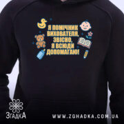 Худі помічник вихователя подарунок, чорного кольору, з капюшоном, на передньому плані видно принт з малюнками і текстом. Фон світлий, що підкреслює контрастність худі.