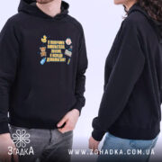 Худі помічник вихователя подарунок, чорного кольору, з капюшоном, на фото видно двох людей, один з яких носить худі. Принт на грудях яскравий, фон світлий.