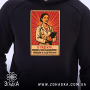 Худі стильний подарунок педіатру, темно-синього кольору, з близького ракурсу, на фоні видно принт з лікарем на червоному фоні, капюшон піднятий, деталі принта чітко видимі, є кишеня-«кенгуру», манжети та пояс з еластичної тканини.