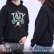 Худі тату майстер, чорного кольору, з капюшоном, з бокового ракурсу, на фоні світлого кольору, з принтом, що містить зображення тату машинок та напис 'TATY МАЙСТЕР', має кишеню-«кенгуру», манжети та пояс з еластичної тканини.