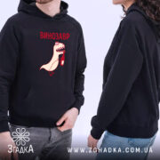 Худі з принтом динозавр, темно-синього кольору, зображене з ракурсу спереду, на фоні світлого простору. Два моделі, один з яких тримає кишеню-«кенгуру», принт з веселим динозавром, що тримає пляшку, виконаний у червоному та світло-кремовому кольорах. Капюшон на обох моделях, манжети та пояс в рибаній текстурі.