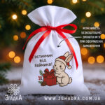 Мішечок подарунок від зайчика з червоною стрічкою на фоні ялинкових вогників