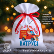 Мішечок від Миколая з іменем Катрусі, білий габардин, червона стрічка, фон — ялинкові вогники.