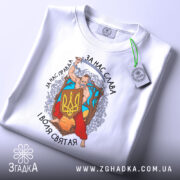 Футболка український козак, білого кольору, зображена зверху, складена на поверхні, фон світло-сірий, принт з козаком, що тримає меч і щит з тризубом, оточений деревами та квітами, текст на футболці: 'За нас слава, за нас правда, і воля святая', на футболці є етикетка з зеленим шнурком.