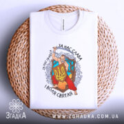 Футболка український козак, білого кольору, зображена зверху, складена на плетеній кошику, фон світло-сірий, принт з козаком, що тримає меч і щит з тризубом, оточений деревами та квітами, текст на футболці: 'За нас слава, за нас правда, і воля святая'.