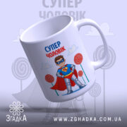 Чашка супер чоловік, біла, з малюнком супергероя, що носить червоний плащ і синій костюм, нахилена вбік, на фоні блакитного неба з хмарами та кольоровими цукерками, ручка кругла, край рівний, глянцева поверхня.