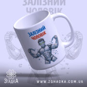 Чашка залізний чоловік, біла з кольоровим принтом, зображення залізного чоловіка з ланцюгами, ручка з правого боку, глянець на поверхні, фон світло-сірий з повторюючим малюнком.