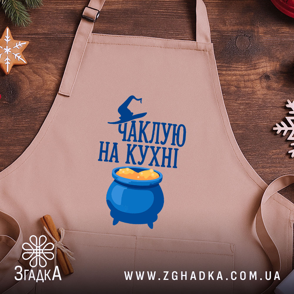 Фартух чаклую на кухні, бежевого кольору, зображений з верхнього ракурсу на дерев'яному фоні, деталі принта включають напис та малюнок казана з варивом, поряд розташовані святкові елементи.