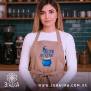 Фартух чаклую на кухні, бежевого кольору, зображений на жінці з фронтального ракурсу, фон кухні з темно-зеленими плитками, принт з написом та казаном з варивом.
