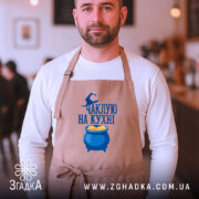 Фартух чаклую на кухні, бежевого кольору, зображений на чоловікові з фронтального ракурсу, фон кафе з розмитими деталями, принт з написом та казаном з варивом.