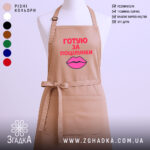 Фартух готую за поцілунки, бежевого кольору, зображений з фронтального ракурсу на манекені, фон світлий, принт з текстом червоного кольору та малюнком губ рожевого кольору.