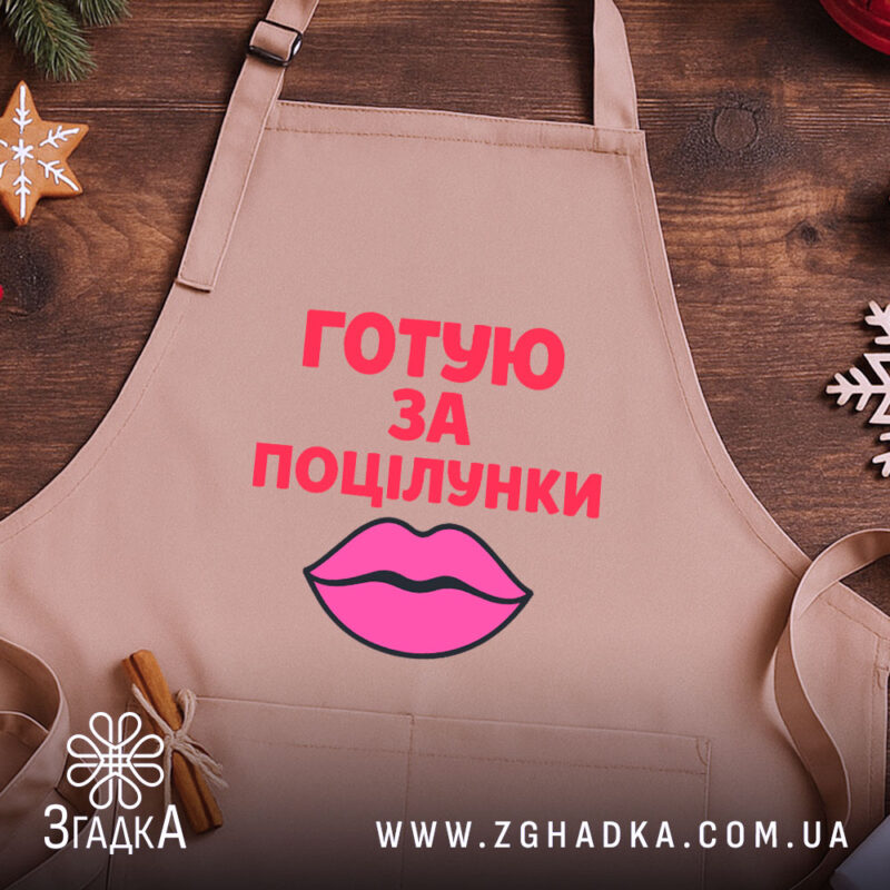 Фартух готую за поцілунки, бежевого кольору, зображений з верхнього ракурсу на дерев'яному фоні, принт з текстом червоного кольору та малюнком губ рожевого кольору, навколо декор у вигляді новорічних прикрас.