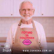 Фартух готую за поцілунки, бежевого кольору, зображений з верхнього ракурсу на дерев'яному фоні, принт з текстом червоного кольору та малюнком губ рожевого кольору, навколо декор у вигляді новорічних прикрас.