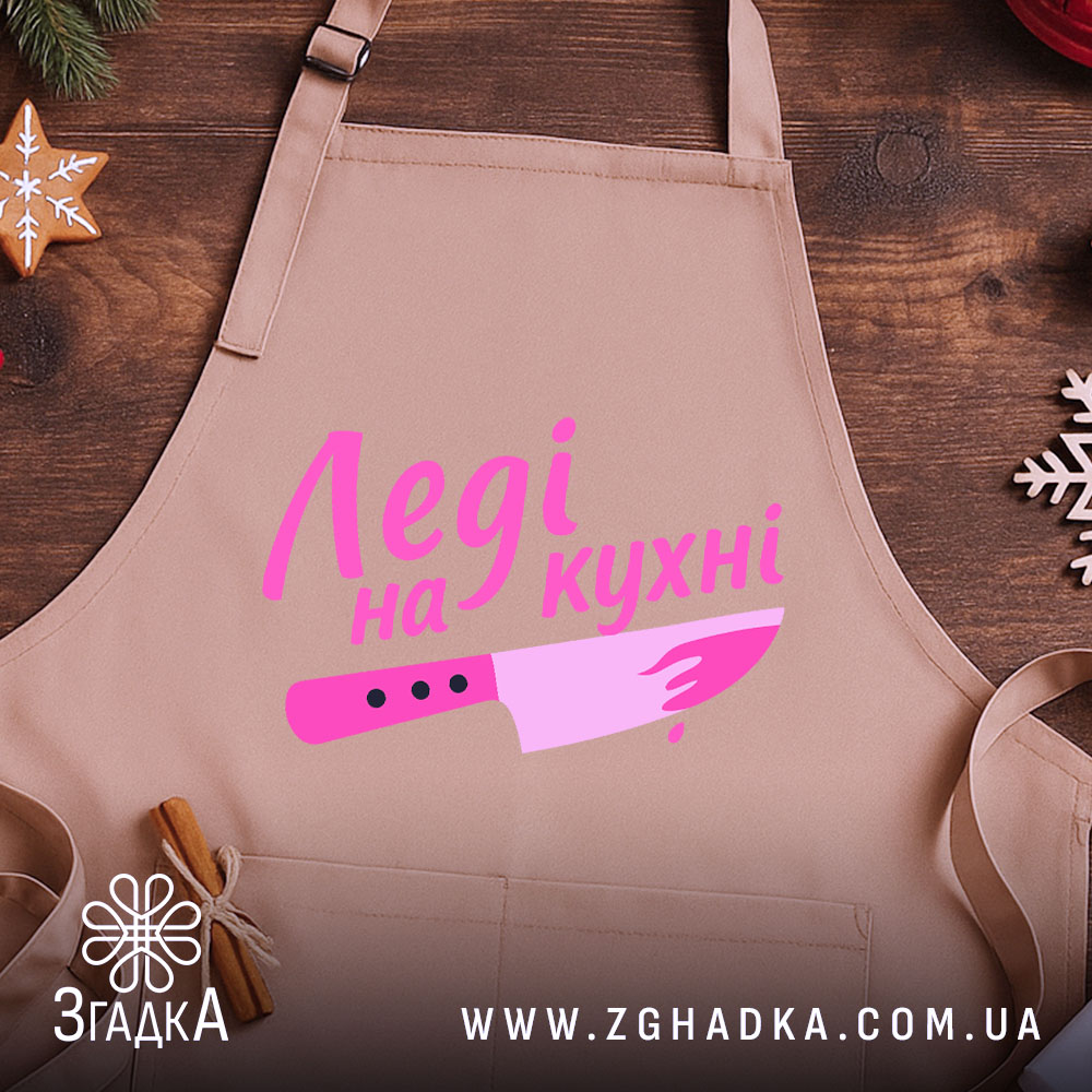 Фартух леді на кухні, бежевого кольору, зображений на дерев'яному фоні, деталі принта включають напис 'Леді на кухні' та малюнок ножа в рожевих відтінках, поруч видно декор у вигляді новорічних зірочок.