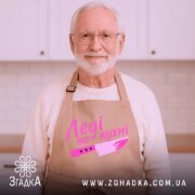 Фартух леді на кухні, бежевого кольору, зображений на дерев'яному фоні, деталі принта включають напис 'Леді на кухні' та малюнок ножа в рожевих відтінках, поруч видно декор у вигляді новорічних зірочок.