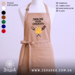 Фартух люблю коли смажать, бежевого кольору, зображений з фронтального ракурсу на манекені, фон світлий, принт з текстом та малюнком вогню і кухонних інструментів.