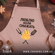 Фартух люблю коли смажать, бежевого кольору, зображений з верхнього ракурсу на дерев'яному фоні, принт з текстом та малюнком вогню і кухонних інструментів, поруч видно новорічні прикраси.