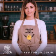 Фартух люблю коли смажать, бежевого кольору, зображений на жінці з фронтального ракурсу, фон кухонний з темно-синьою плиткою, принт з текстом та малюнком вогню і кухонних інструментів.
