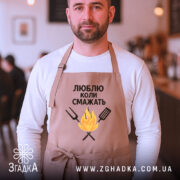 Фартух люблю коли смажать, бежевого кольору, зображений на чоловікові з фронтального ракурсу, фон кафе з розмитими фігурами людей, принт з текстом та малюнком вогню і кухонних інструментів.