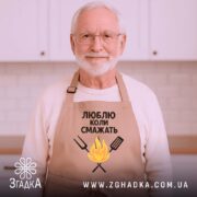 Фартух люблю коли смажать, бежевого кольору, зображений з верхнього ракурсу на дерев'яному фоні, принт з текстом та малюнком вогню і кухонних інструментів, поруч видно новорічні прикраси.