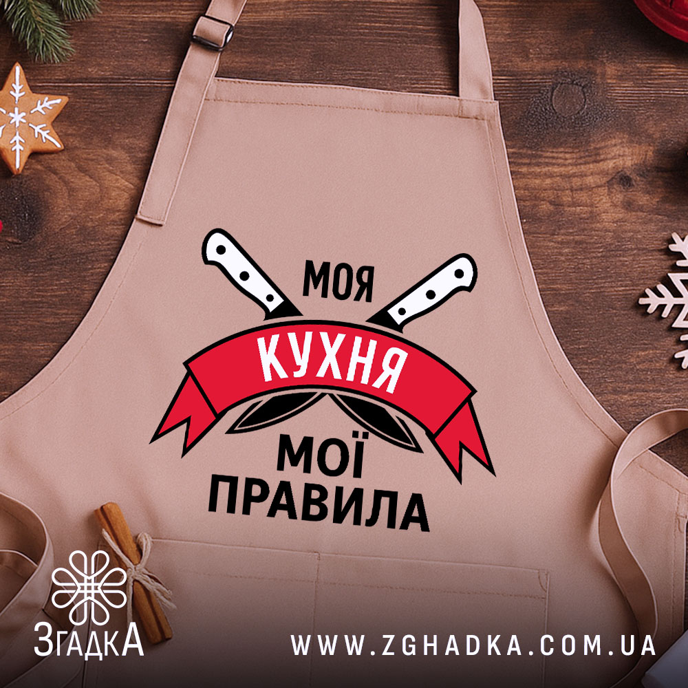 Фартух моя кухня мої правила. Бежевий фартух, знятий з близького ракурсу на дерев'яному фоні. На фоні видно декор у вигляді новорічних елементів. Принт з написом 'Моя кухня мої правила' та ножами.