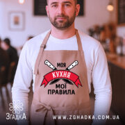 Фартух моя кухня мої правила. Бежевий фартух, зображений на чоловікові, який стоїть у кафе. Фон розмитий, з видимими елементами інтер'єру. Принт містить напис 'Моя кухня мої правила' на червоному банері з ножами.