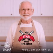 Фартух моя кухня мої правила. Бежевий фартух, знятий з близького ракурсу на дерев'яному фоні. На фоні видно декор у вигляді новорічних елементів. Принт з написом 'Моя кухня мої правила' та ножами.